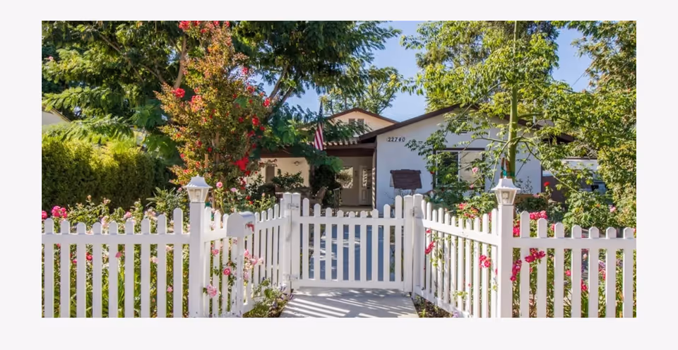 Front of a single-story house with a white picket fence and a garden-lined path to the entrance.