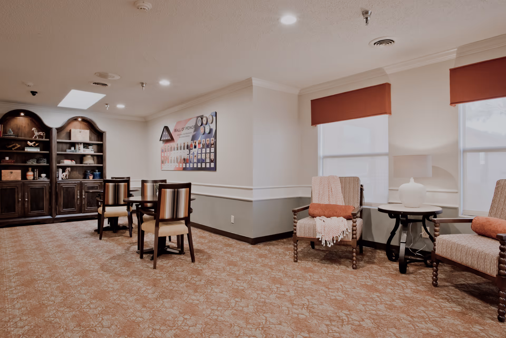 A cozy common area in a nursing and rehabilitation center featuring a round table with four chairs, two armchairs with orange cushions and a throw blanket, a side table with a lamp, a large wooden cabinet with shelves holding decorative items, and a 'Wall of Honor' display on the wall.