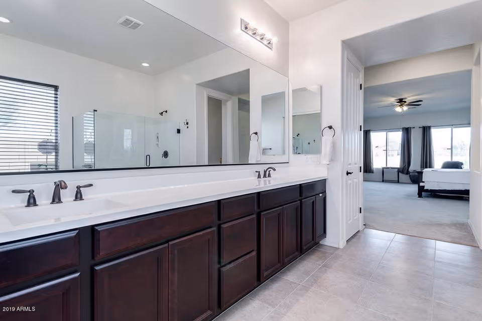 A modern bathroom with a long white countertop featuring two sinks and dark wooden cabinets underneath. Above the countertop is a large mirror with lights mounted on the wall. To the left, there is a glass-enclosed shower with a window beside it. The bathroom floor is tiled, and through an open doorway, a bedroom with a ceiling fan, large windows, and a bed is visible.