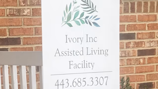 Vertical sign on brick exterior reads Ivory Inc Assisted Living Facility with phone number; bench nearby.