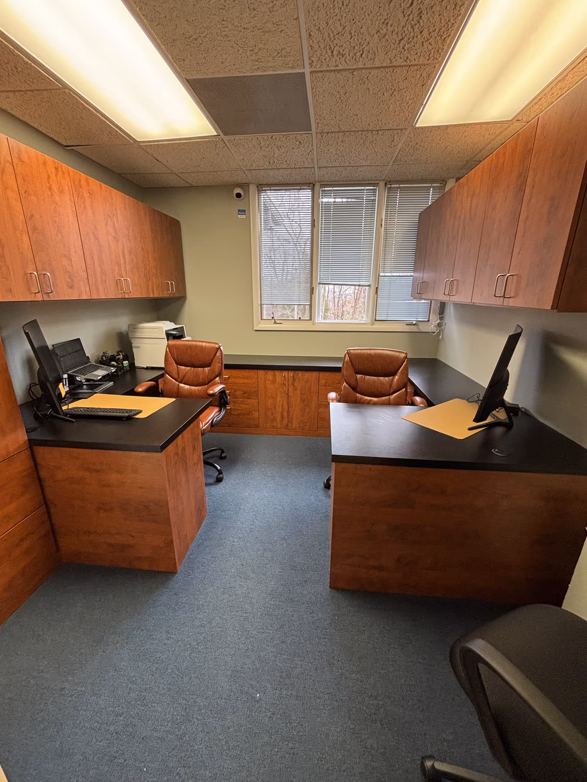 Two wooden desks face each other in a small office with cabinets and a bright window.