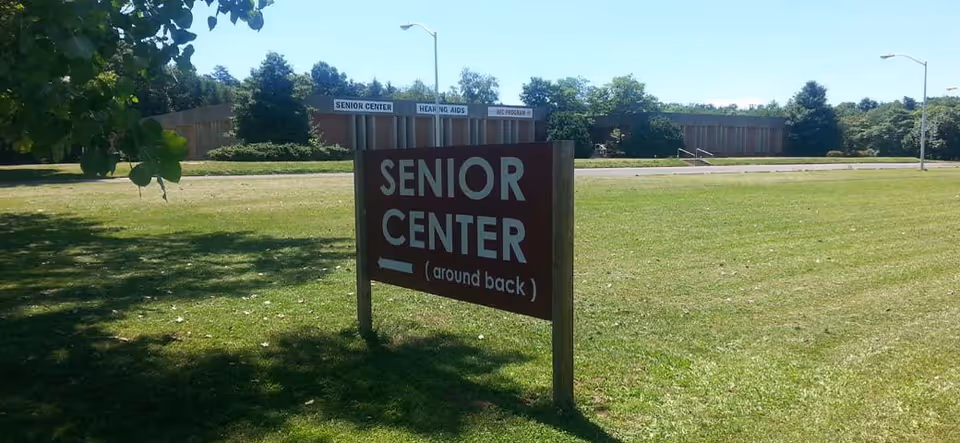 Red wooden sign reading Senior Center on a grassy lawn with a building in background.