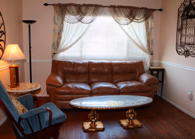 A cozy living room with a brown leather sofa centered under a window with sheer curtains and a decorative valance. In front of the sofa is an oval marble coffee table with ornate gold legs. To the left is a blue upholstered rocking chair next to a round side table with a lamp. The room has wooden flooring and light-colored walls with a decorative metal wall hanging on the right.