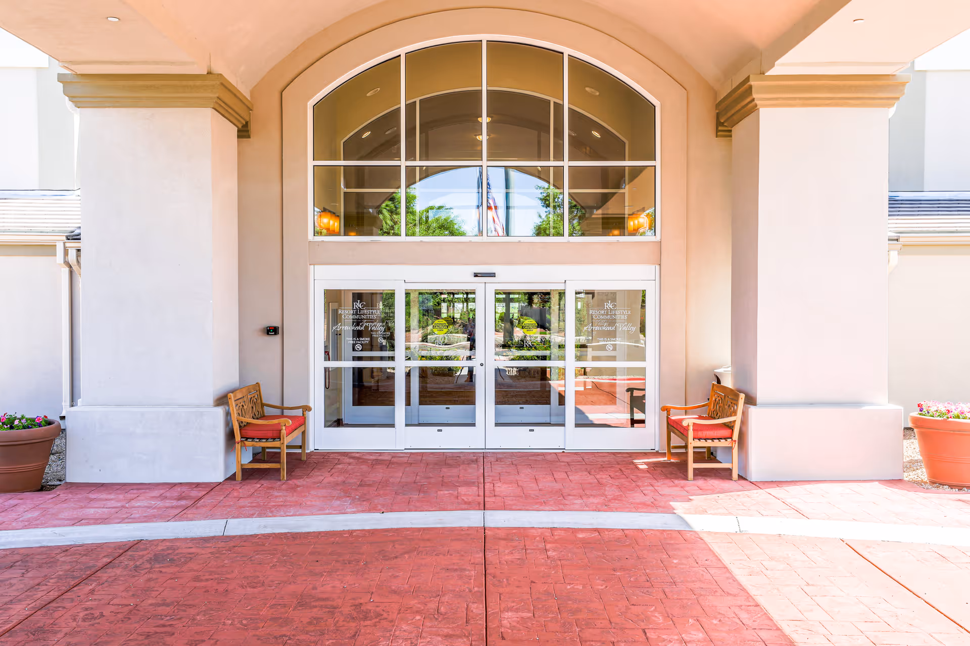 Entrance of Arrowhead Valley Retirement Resort featuring glass double doors under a large arched window, flanked by two columns with wooden benches with red cushions on either side and large flower pots.
