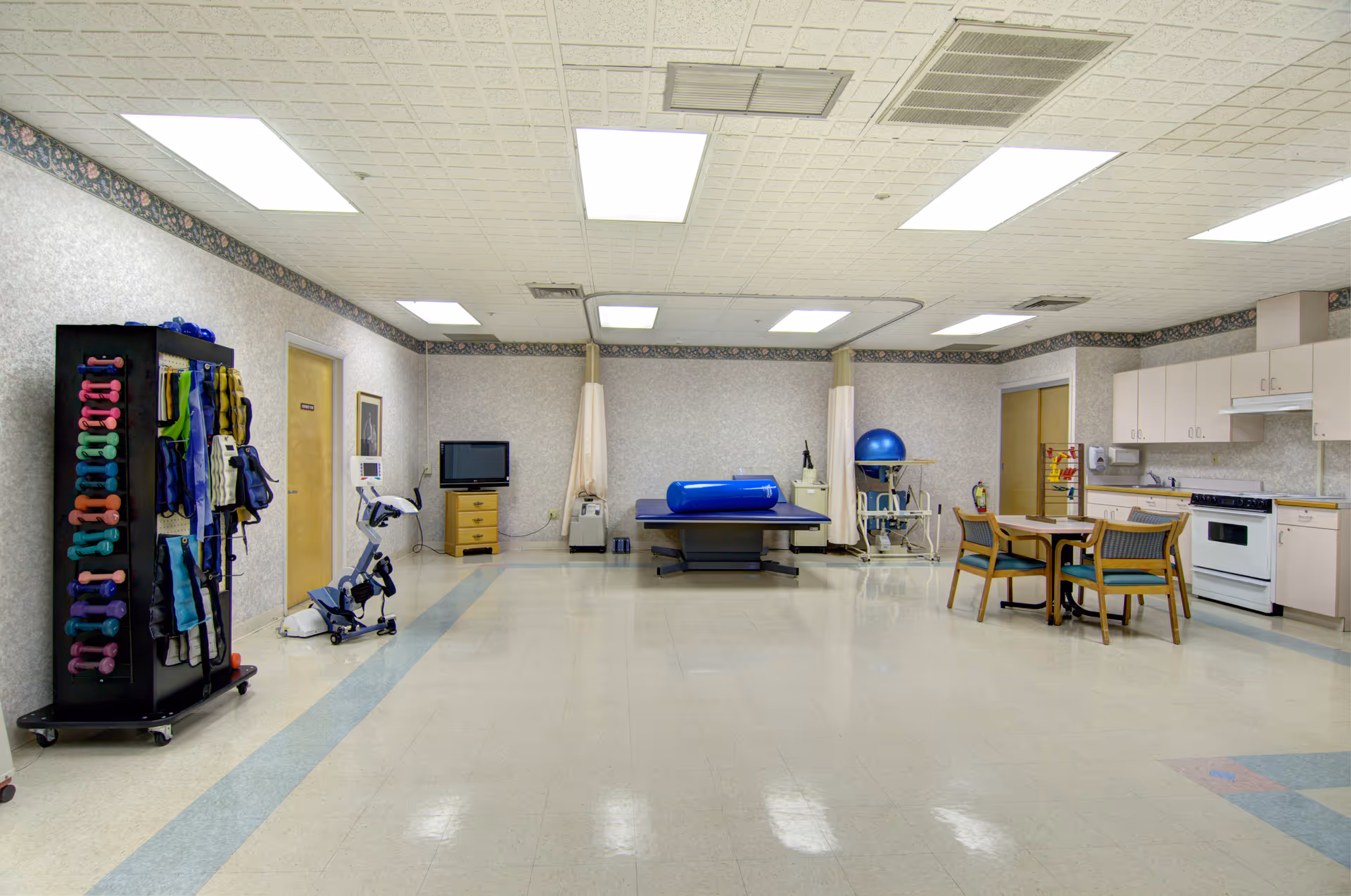 Indoor rehabilitation gym with exercise equipment, a table, and a kitchenette.