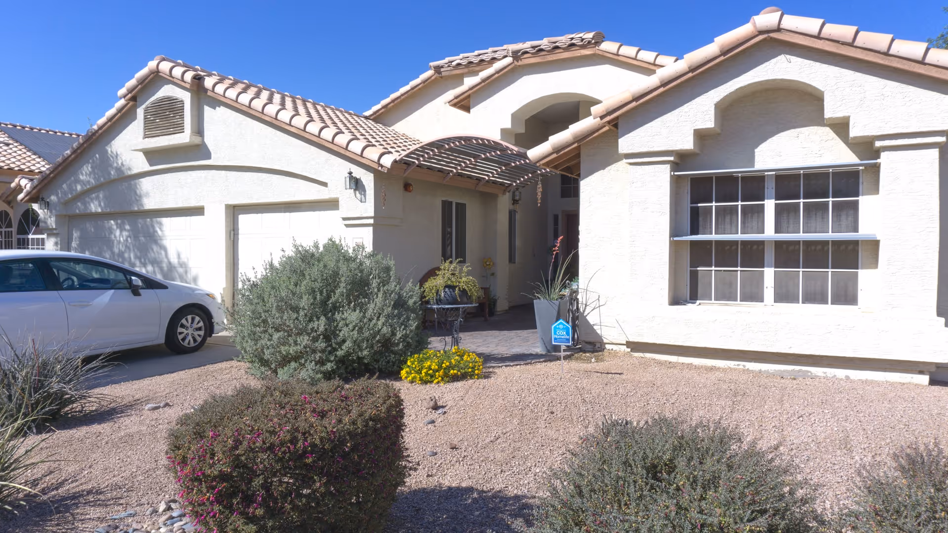 Front exterior view of a single-story house with a tiled roof, a driveway with a white car parked, desert landscaping with bushes and gravel, and a covered entrance with a small table and chairs.