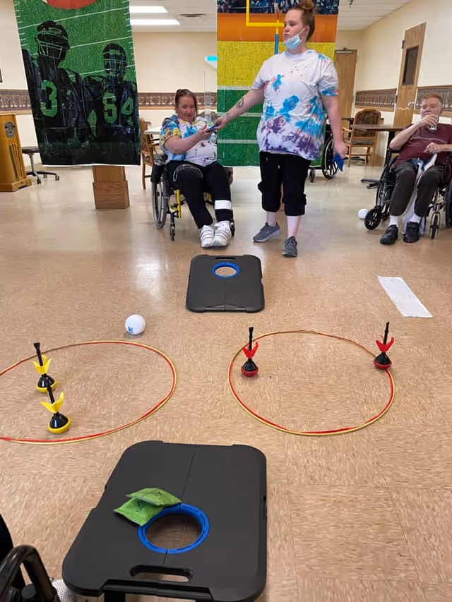 Three people in a senior living facility common area playing an indoor game involving bean bags and rings on the floor. Two individuals are seated in wheelchairs, and one person is standing, handing a bean bag to a seated player. The room has a linoleum floor and sports-themed banners hanging on the walls.