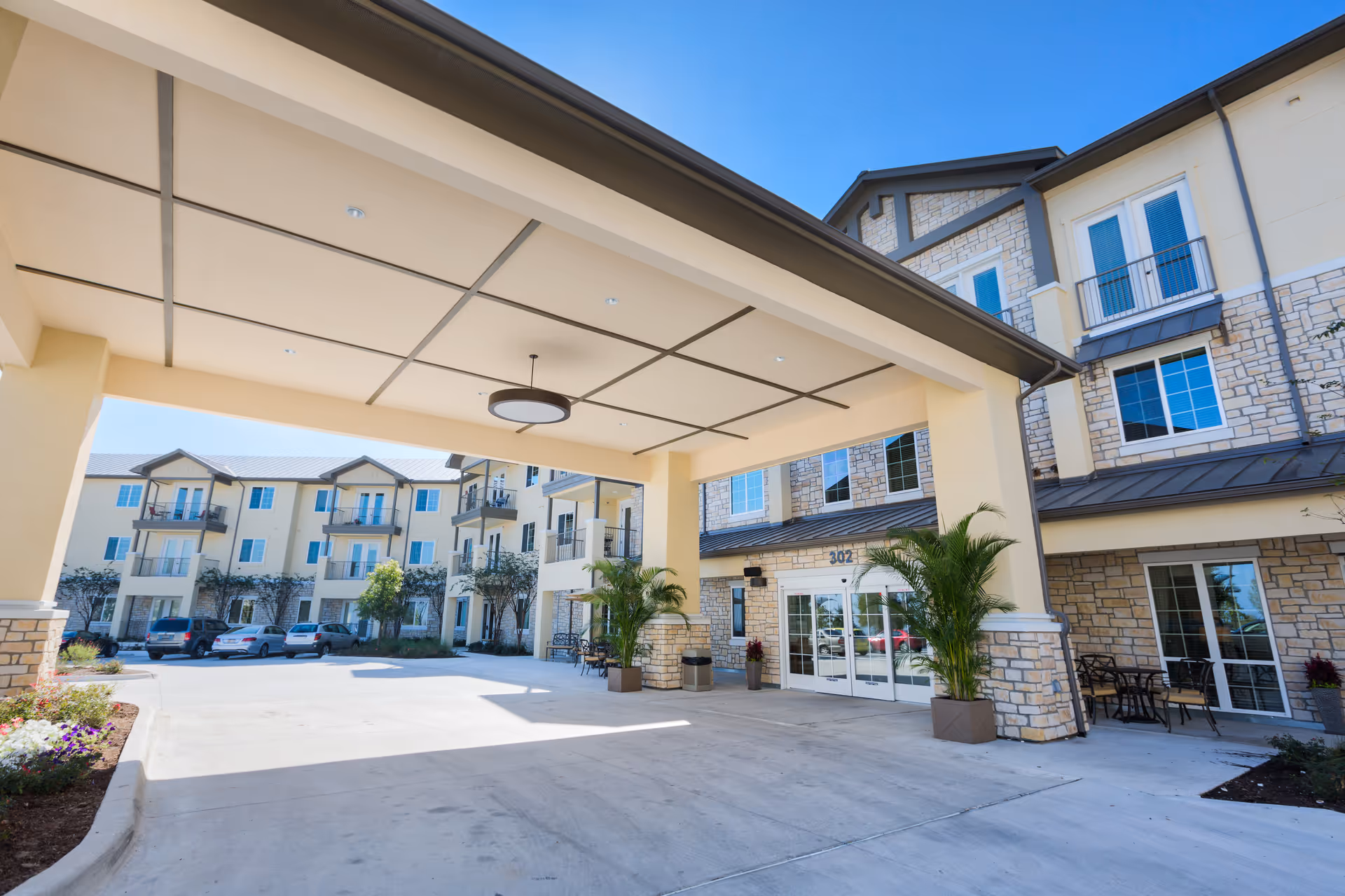 Covered porte-cochere entrance of a multi-story senior living building with balconies, potted plants, and parked cars.
