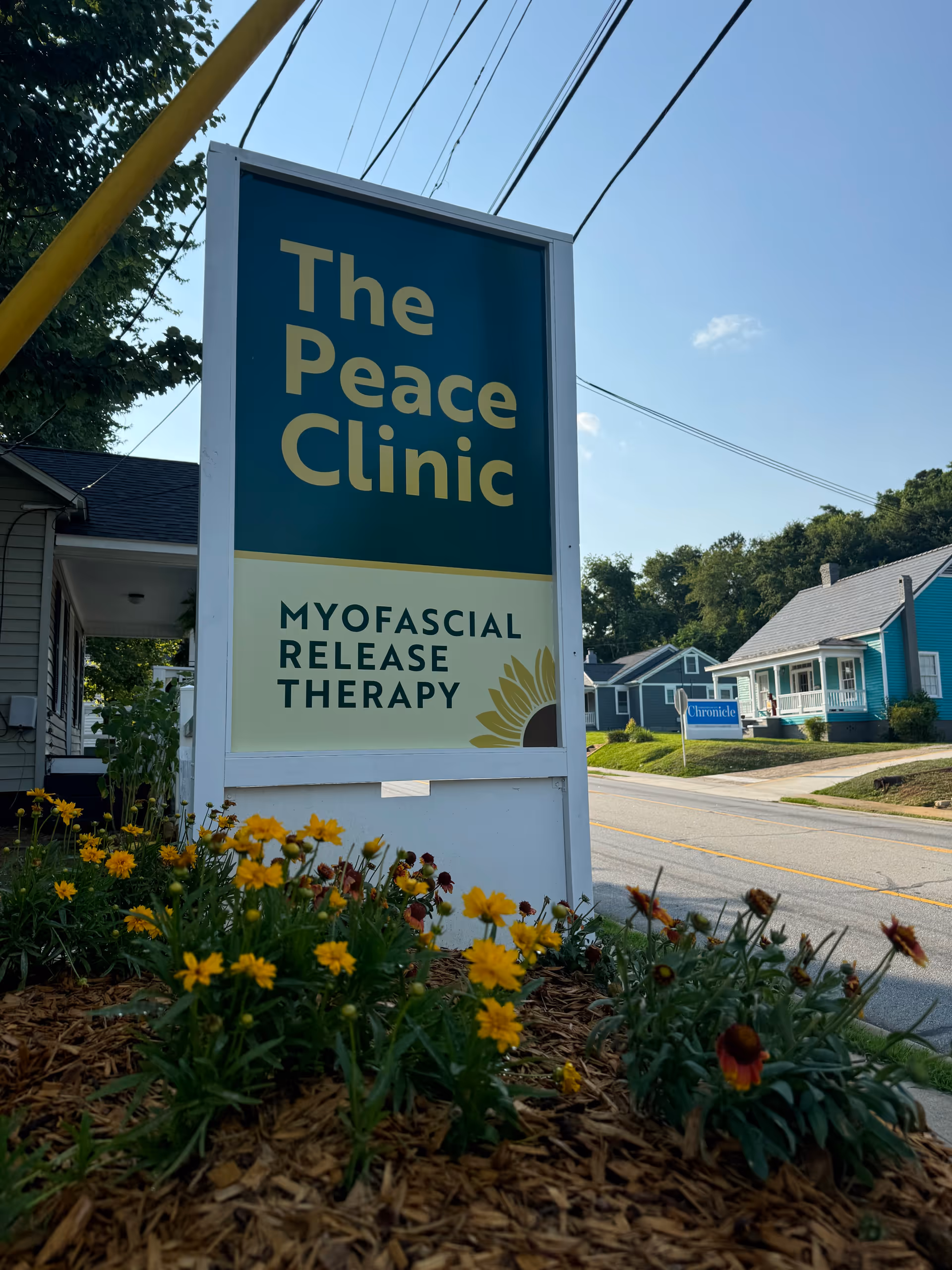Tall outdoor sign reading The Peace Clinic with Myofascial Release Therapy beside a bed of flowers.
