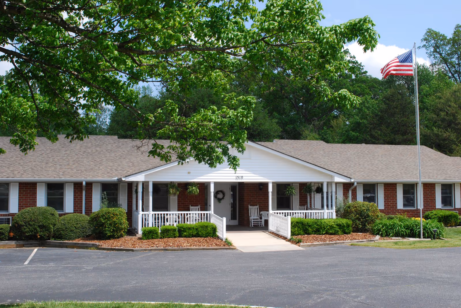 Front exterior view of a single-story brick building with a white porch, rocking chairs, hanging plants, and an American flag on a flagpole. The building is surrounded by green bushes and trees under a partly cloudy sky.