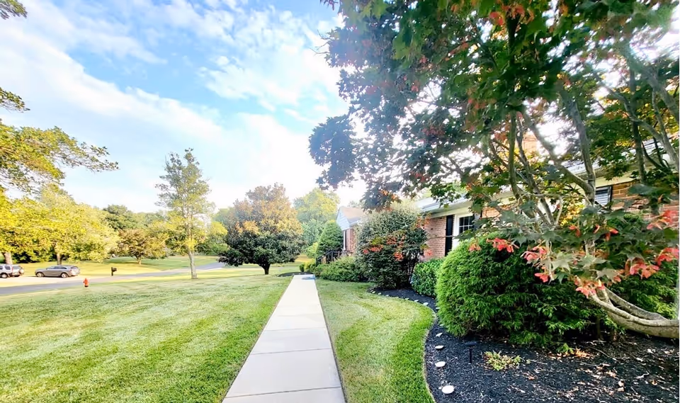 A sunny outdoor scene showing a paved walkway leading to a brick building partially visible on the right, surrounded by well-maintained green grass, bushes, and trees with some leaves turning red. Several cars are parked along the street in the background under a partly cloudy sky.