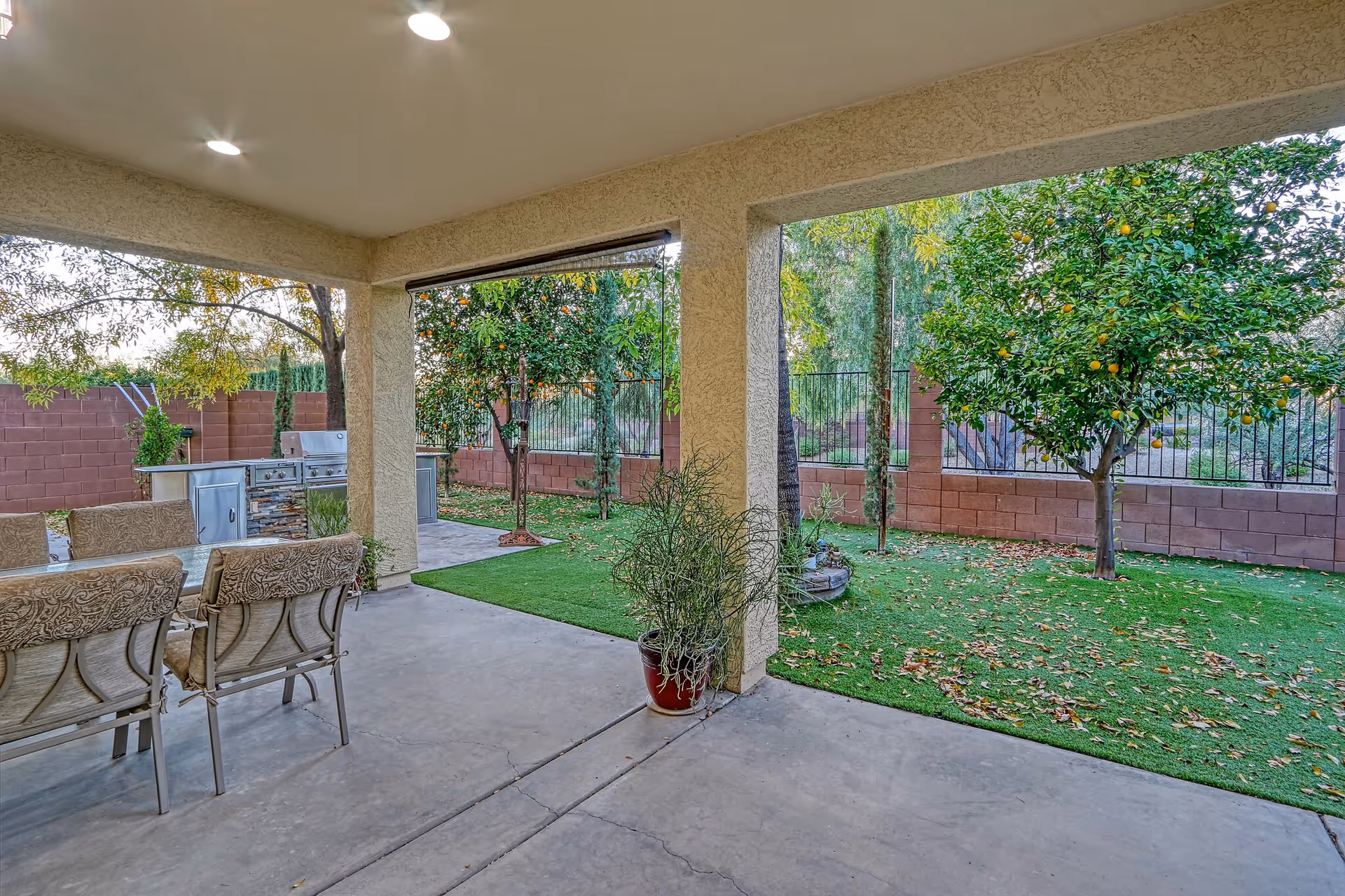 Covered outdoor patio area with cushioned chairs around a table, a built-in outdoor grill, and a view of a fenced backyard with green grass, trees, and plants.