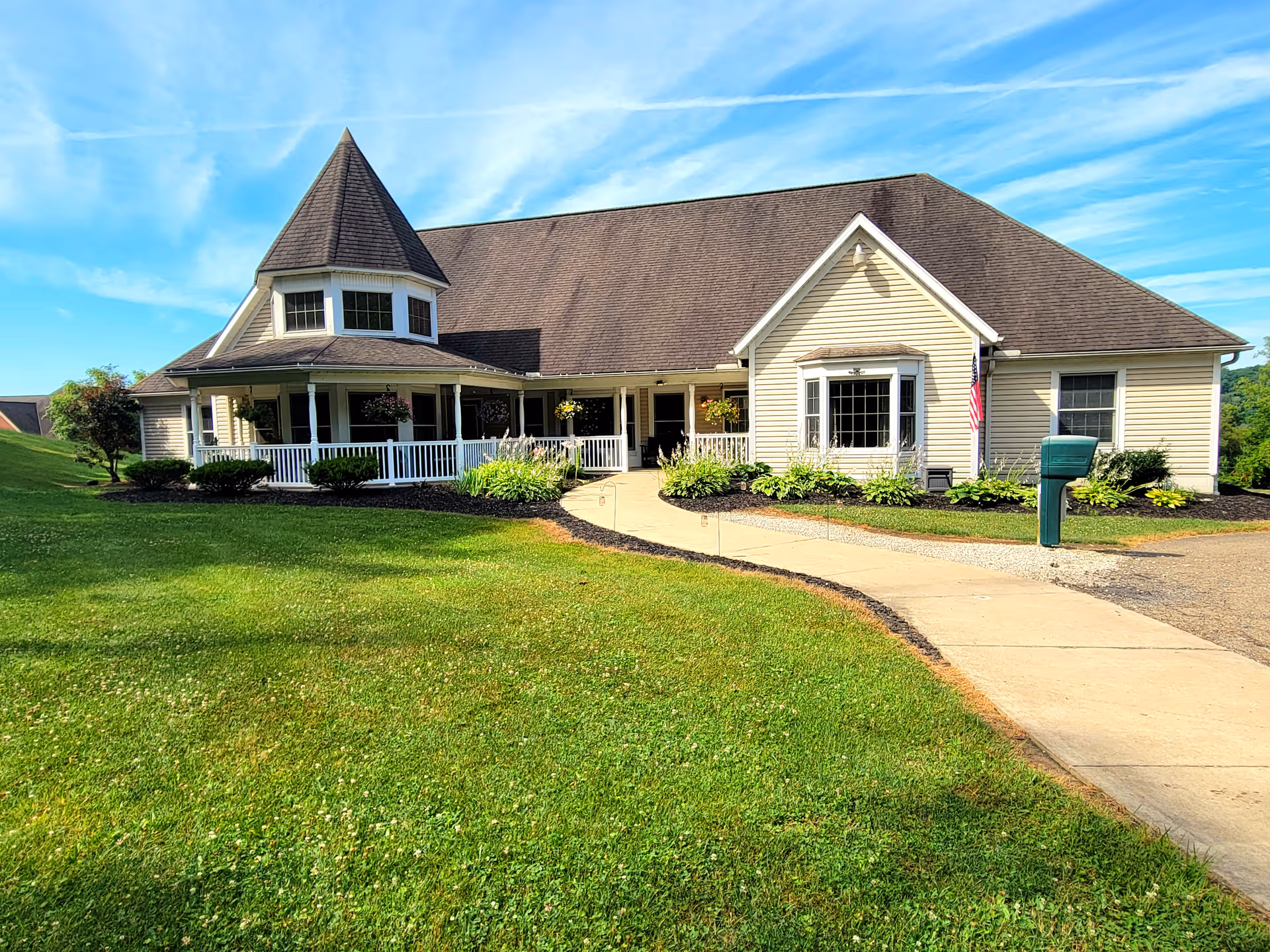 Exterior view of a single-story building with beige siding and a steep roof, featuring a turret-like structure with windows and a wraparound porch with white railings. The building is surrounded by a well-maintained lawn, shrubs, and a concrete walkway leading to the entrance under a bright blue sky with some clouds.