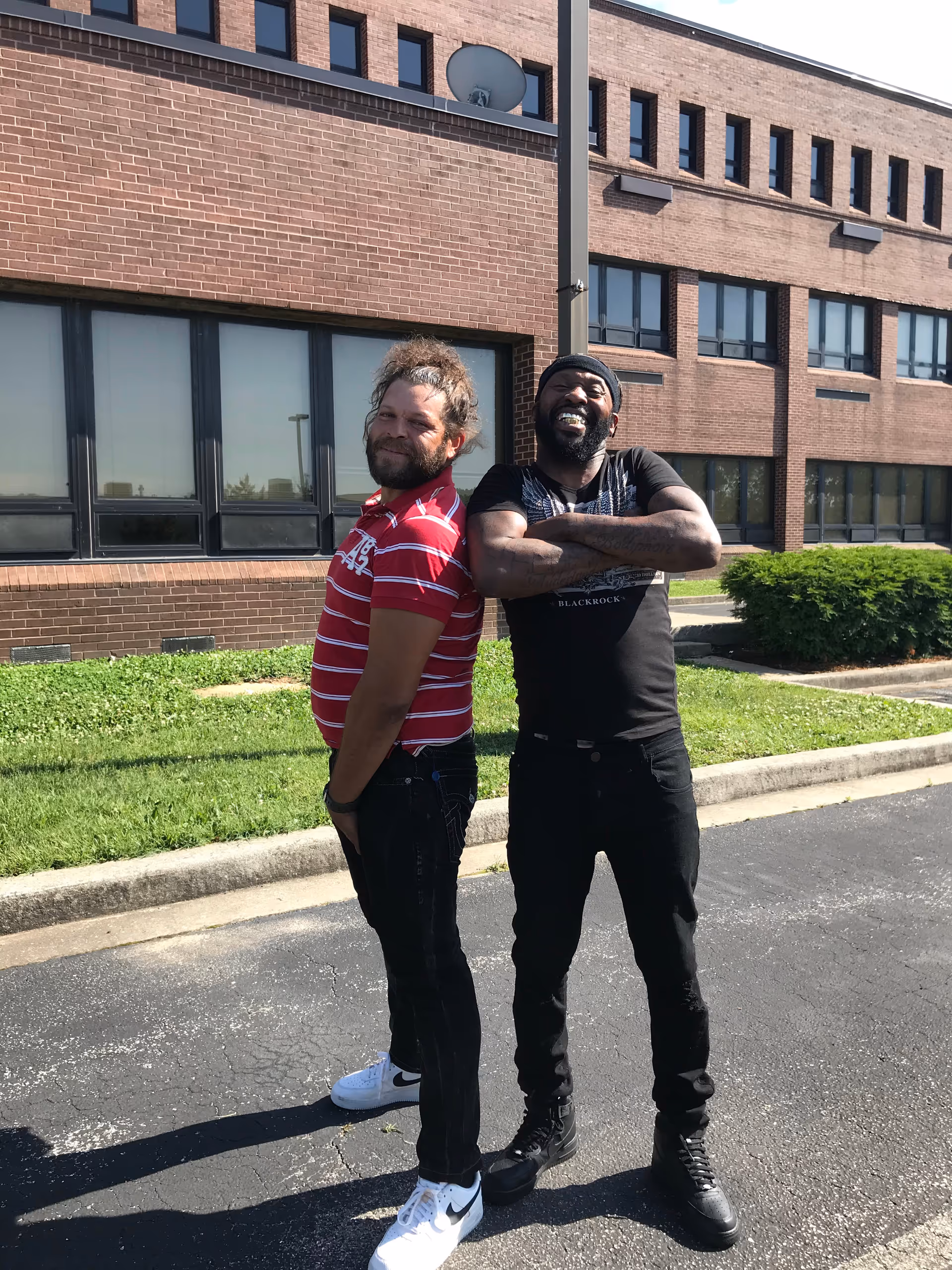 Two men standing outside a brick building, posing and smiling.