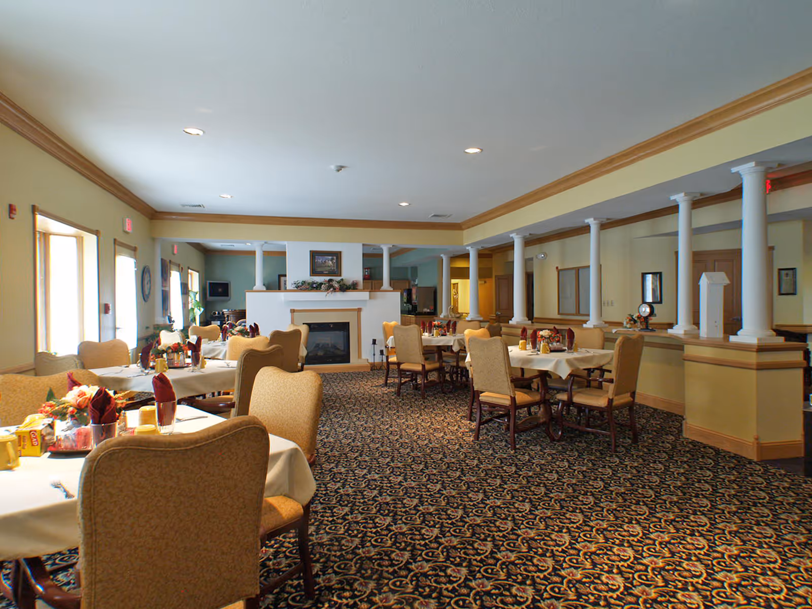 Dining room with multiple set tables and upholstered chairs, a central fireplace, and decorative white columns.