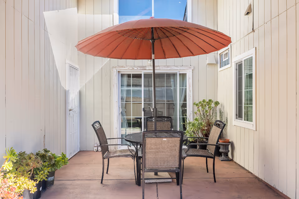 Small enclosed patio with a round table, four chairs, a red umbrella, potted plants, and sliding glass doors to the building.