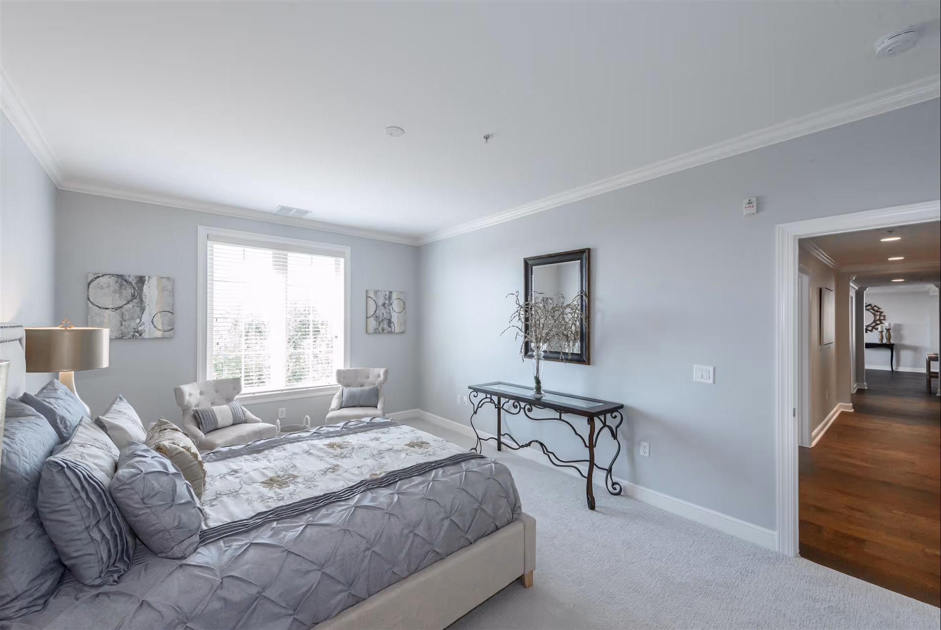 A bright bedroom with a large bed covered in gray and floral bedding, two beige armchairs with pillows near a window with white blinds, a decorative console table with a vase and dried branches, and a mirror hanging above it. The walls are painted light gray and the floor is carpeted in white. An open doorway leads to a hallway with wooden flooring.