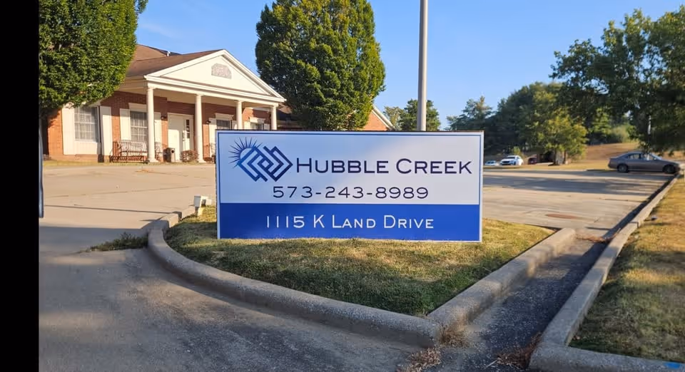 A freestanding sign reading "Hubble Creek" with phone number and address in front of a brick building with columns and a parking lot.