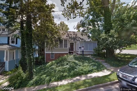 Front of a single-story house with a screened porch, overgrown greenery and trees, and a person standing on the porch.