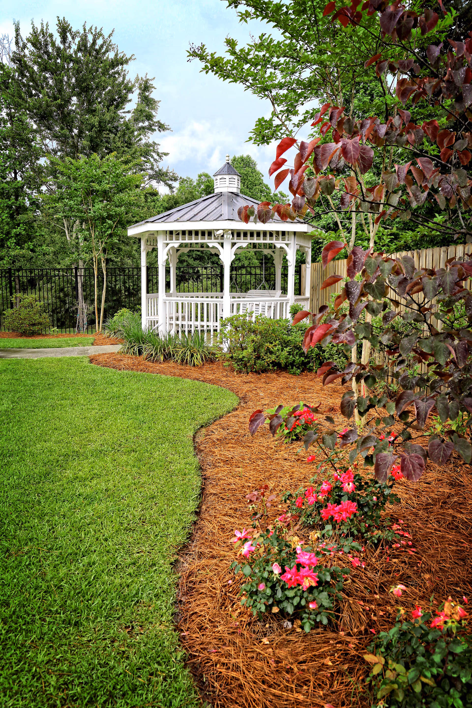 A well-maintained garden area featuring a white gazebo with a metal roof surrounded by green grass, trees, shrubs, and blooming pink flowers along a mulched flower bed.