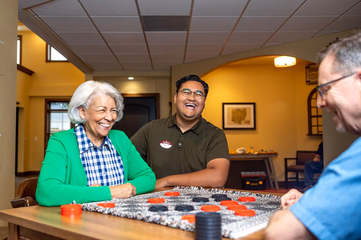Three people sitting around a table playing a board game with red and black pieces, smiling and laughing together in a warmly lit room with yellow walls and framed artwork.