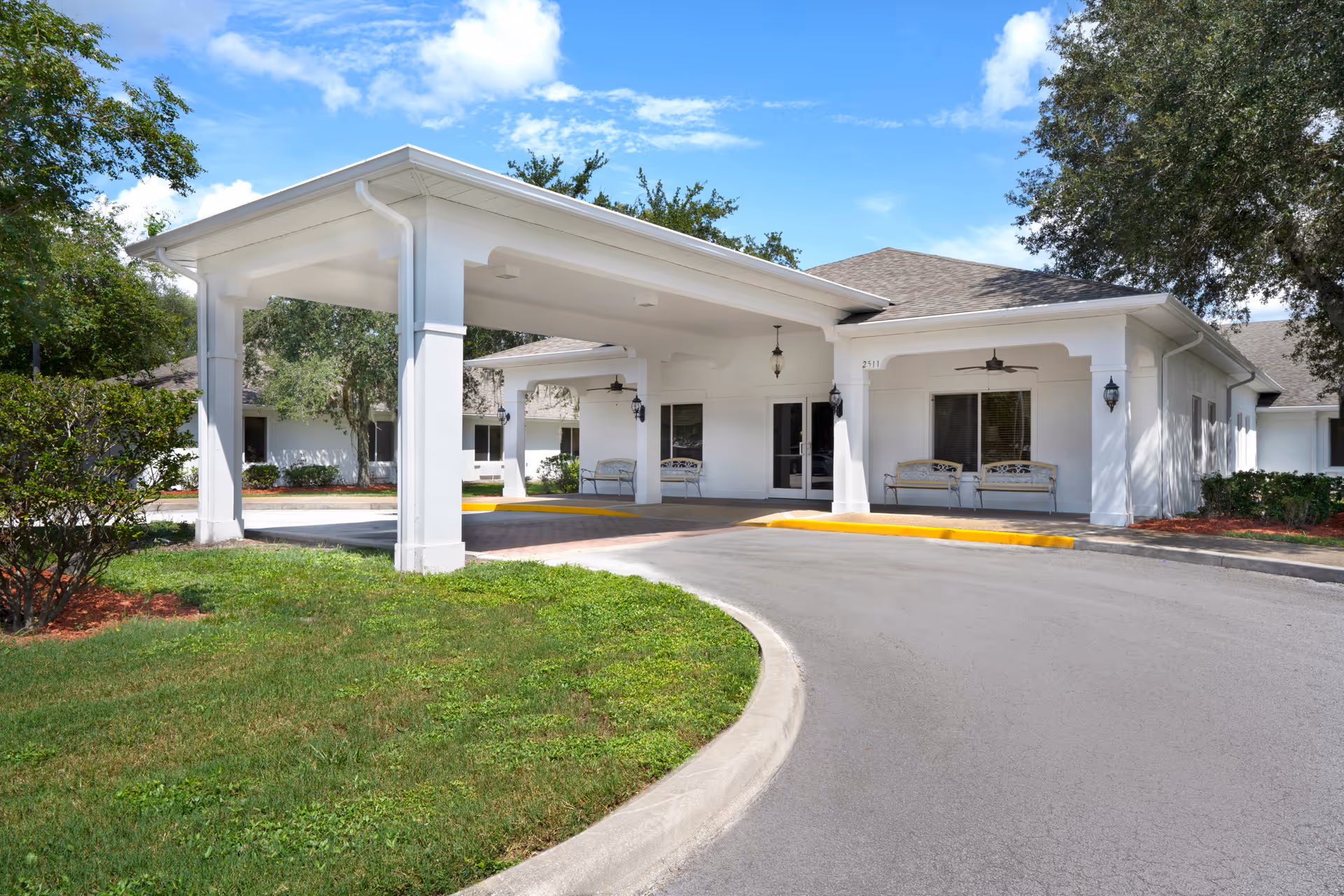 Exterior view of Harborview Health Center Kissimmee showing a covered entrance with white pillars, benches, and a curved driveway surrounded by green grass and trees under a blue sky with some clouds.