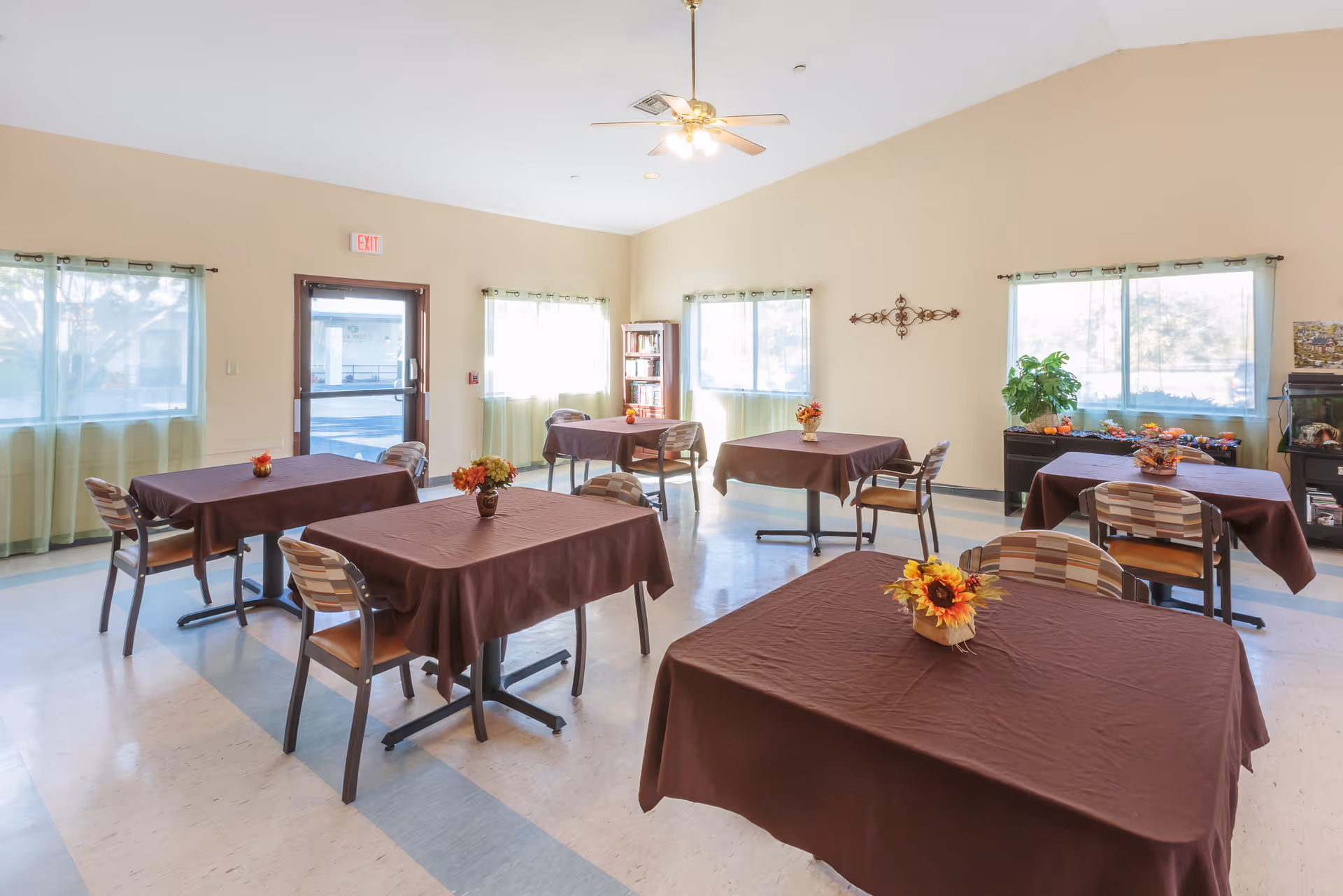 A bright dining room with several tables covered in brown tablecloths, each decorated with a small floral centerpiece. The room has large windows with light green curtains, a ceiling fan with lights, and a bookshelf in the corner. There is an exit door and some plants and decorations along the walls.