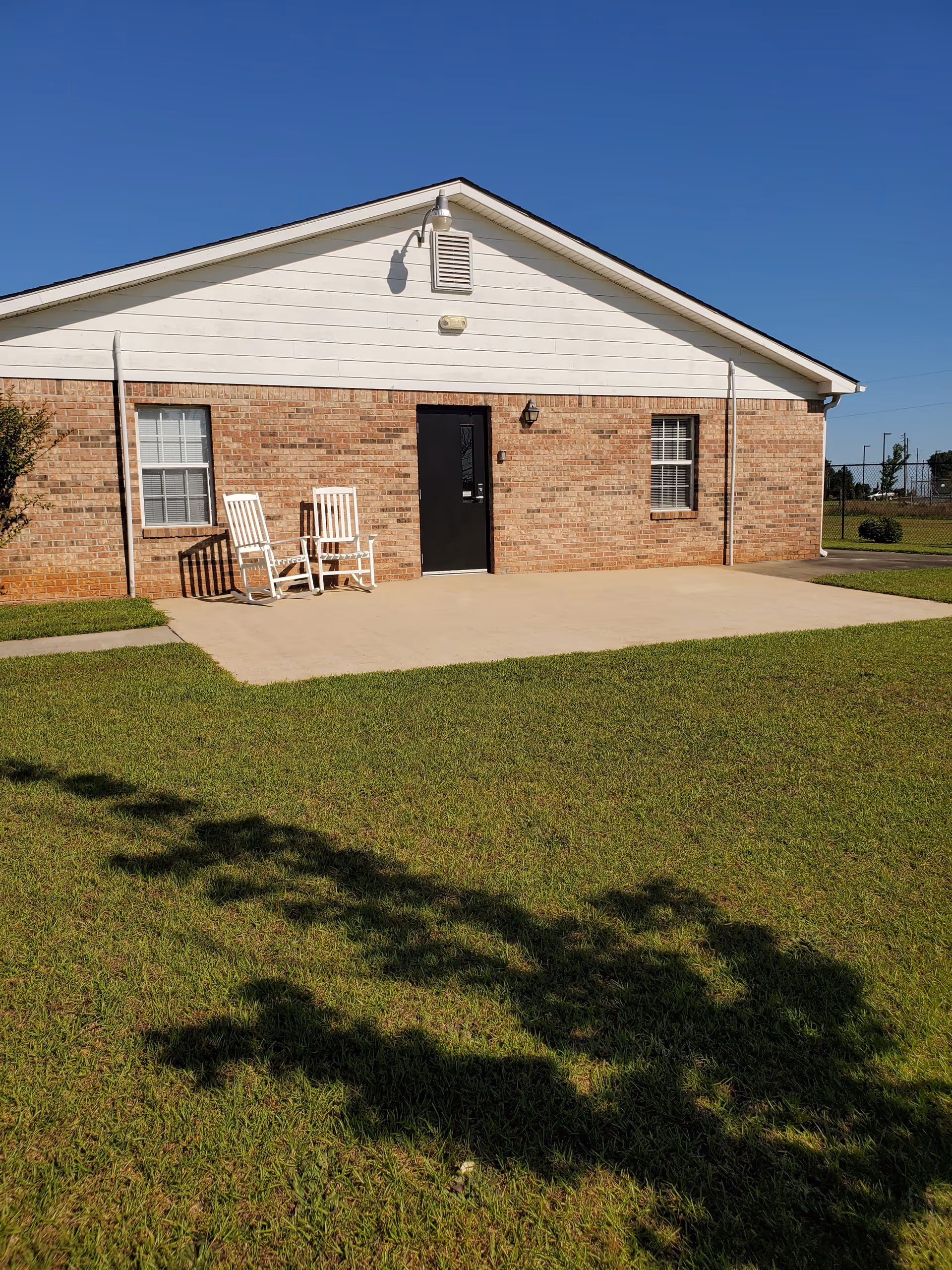 Exterior view of a single-story brick building with a white gable roof. There are two white rocking chairs on a concrete patio in front of a black door, flanked by two windows with white frames. The building is surrounded by a well-maintained green lawn under a clear blue sky.