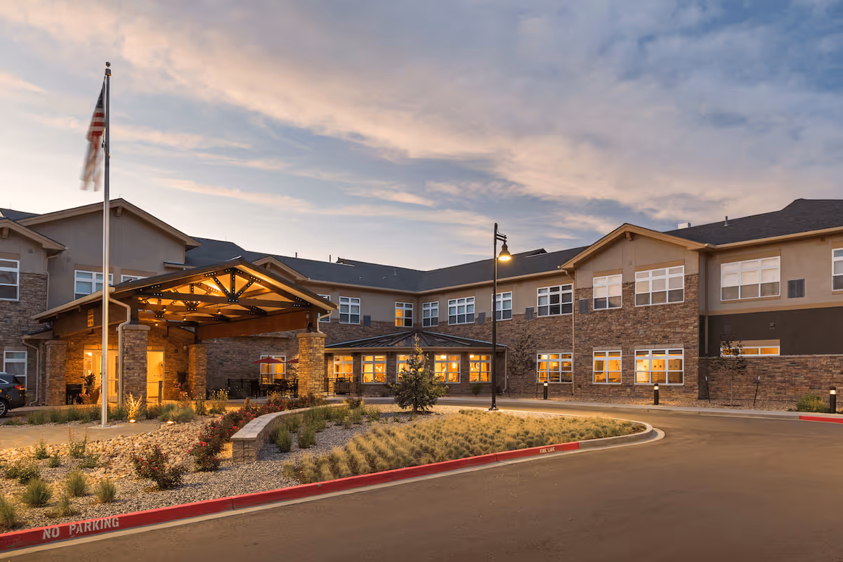 Front exterior of a two-story senior living building at dusk with a covered entrance, landscaped circular drive, and lit windows.