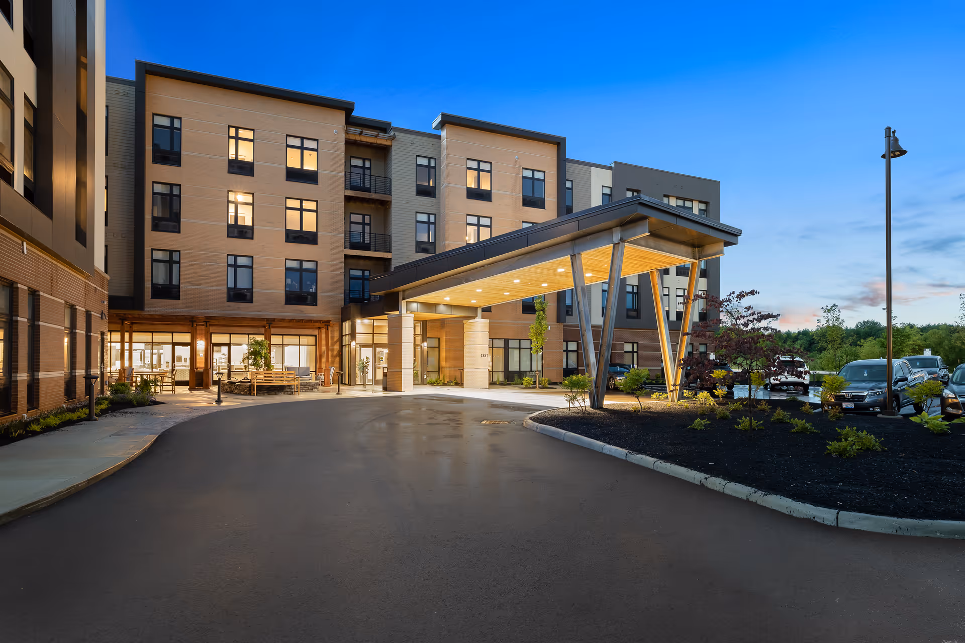 Exterior view of a modern multi-story senior living facility named The Summit of Blue Ash during dusk, showing the entrance with a covered drop-off area, well-lit windows, landscaped garden beds, and parked cars.