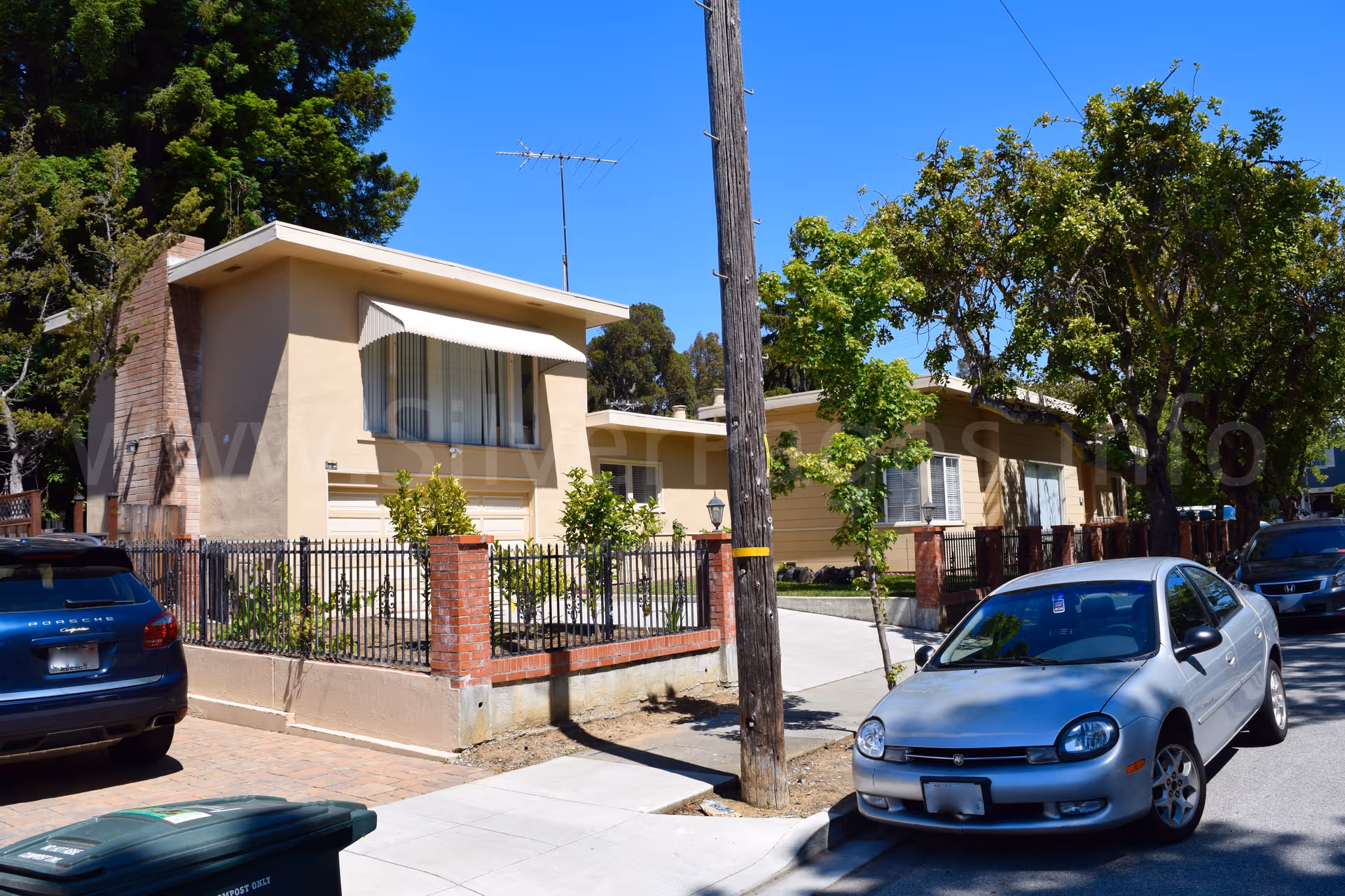 Single-story beige residential care home with brick fence, trees, and parked cars along the street.