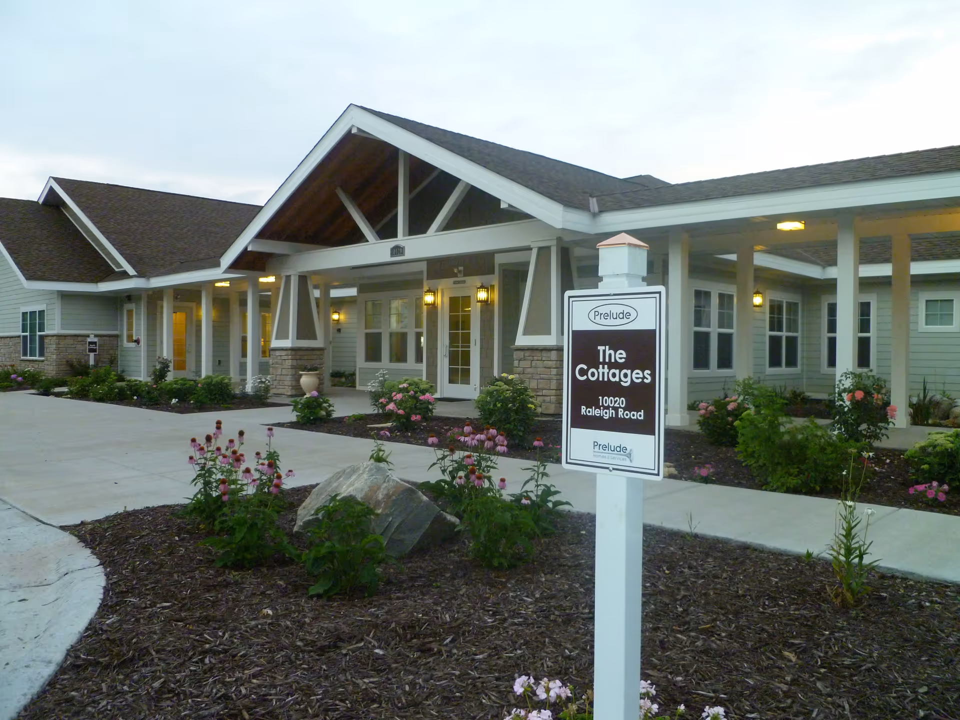 Exterior view of a single-story senior living facility named The Cottages at Prelude Homes & Services, featuring a covered entrance with pillars, landscaped flower beds, and a signpost with the address 10020 Raleigh Road.