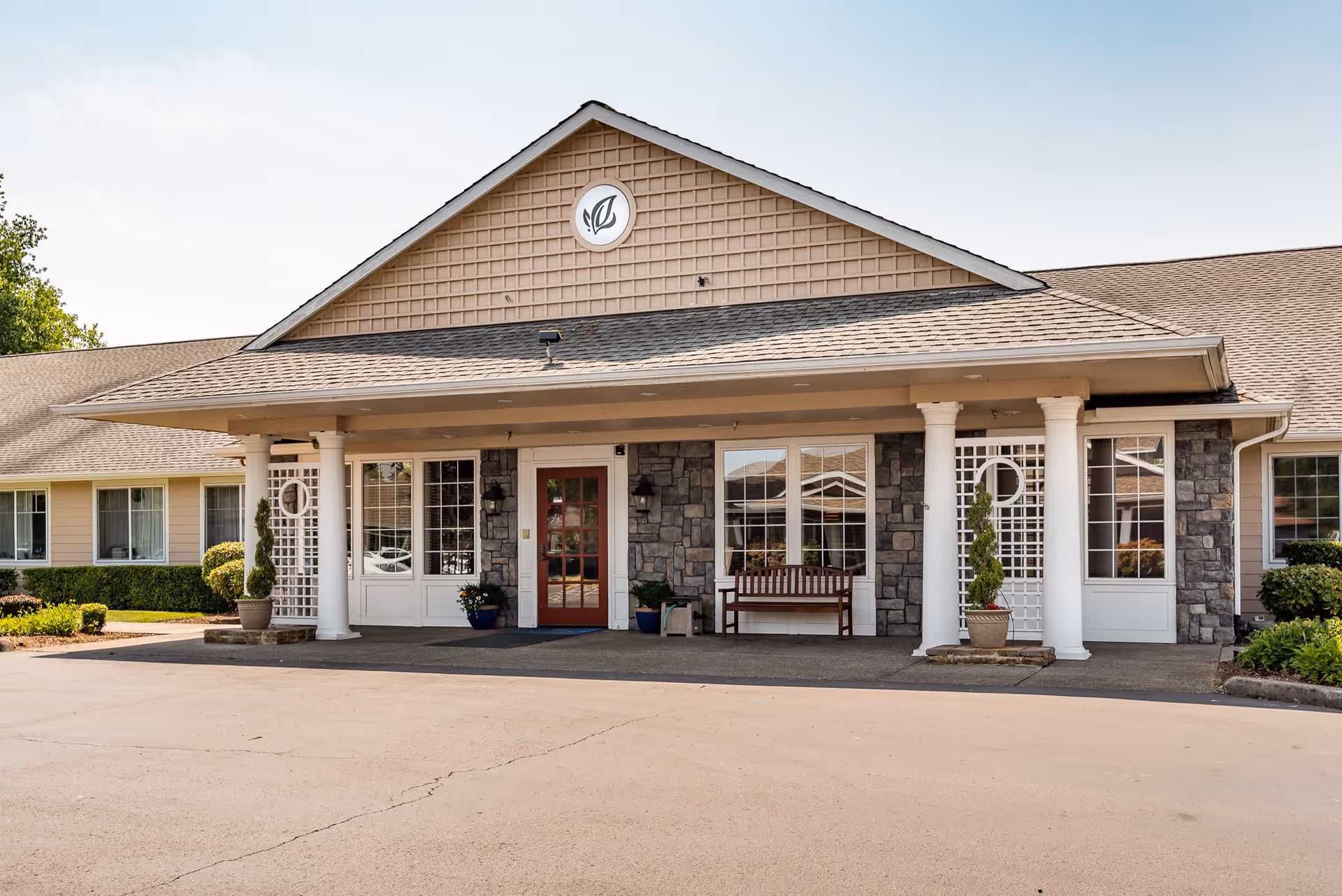 Front exterior view of McMinnville Memory Care facility showing a single-story building with a covered entrance supported by white columns, stone facade around the entrance, large windows, a bench, potted plants, and a circular emblem with a leaf design above the entrance.