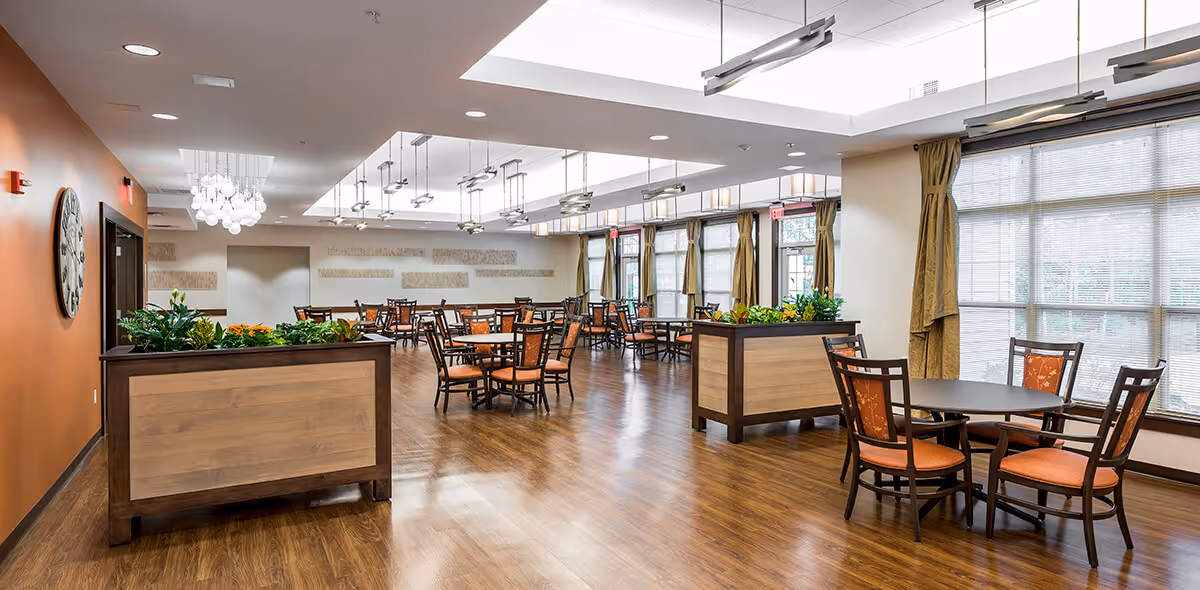 A spacious dining room with multiple round tables and chairs arranged neatly. The room features large windows with curtains allowing natural light to fill the space. There are wooden floorings and planters with green plants adding a touch of nature. Modern ceiling lights hang above the tables, and a large wall clock is visible on an orange accent wall.