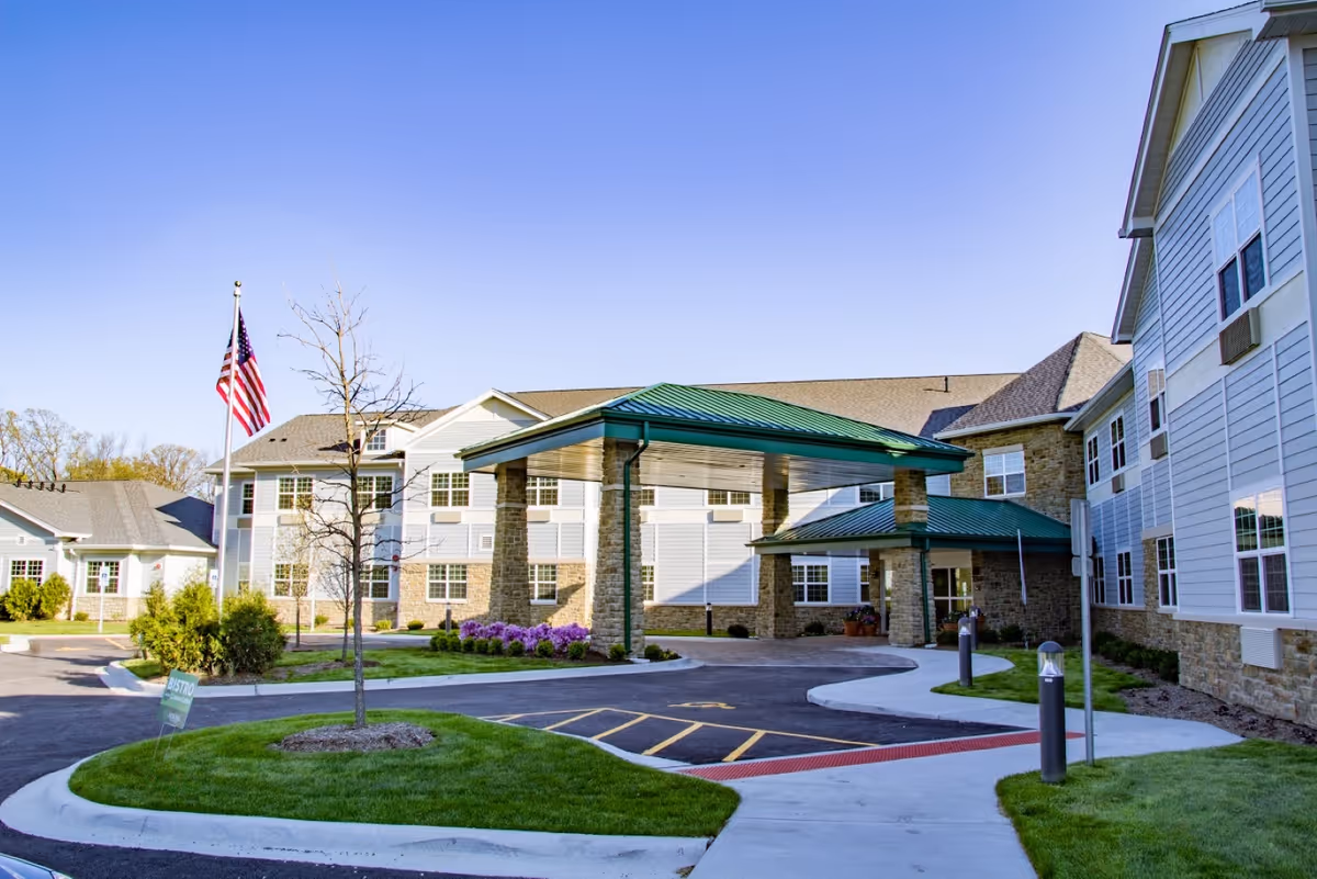 Exterior view of New Perspective Senior Living | Long Grove building with a covered entrance, green metal roof, stone pillars, and an American flag on a flagpole. The sky is clear and blue, and there is a well-maintained lawn and paved driveway in front.