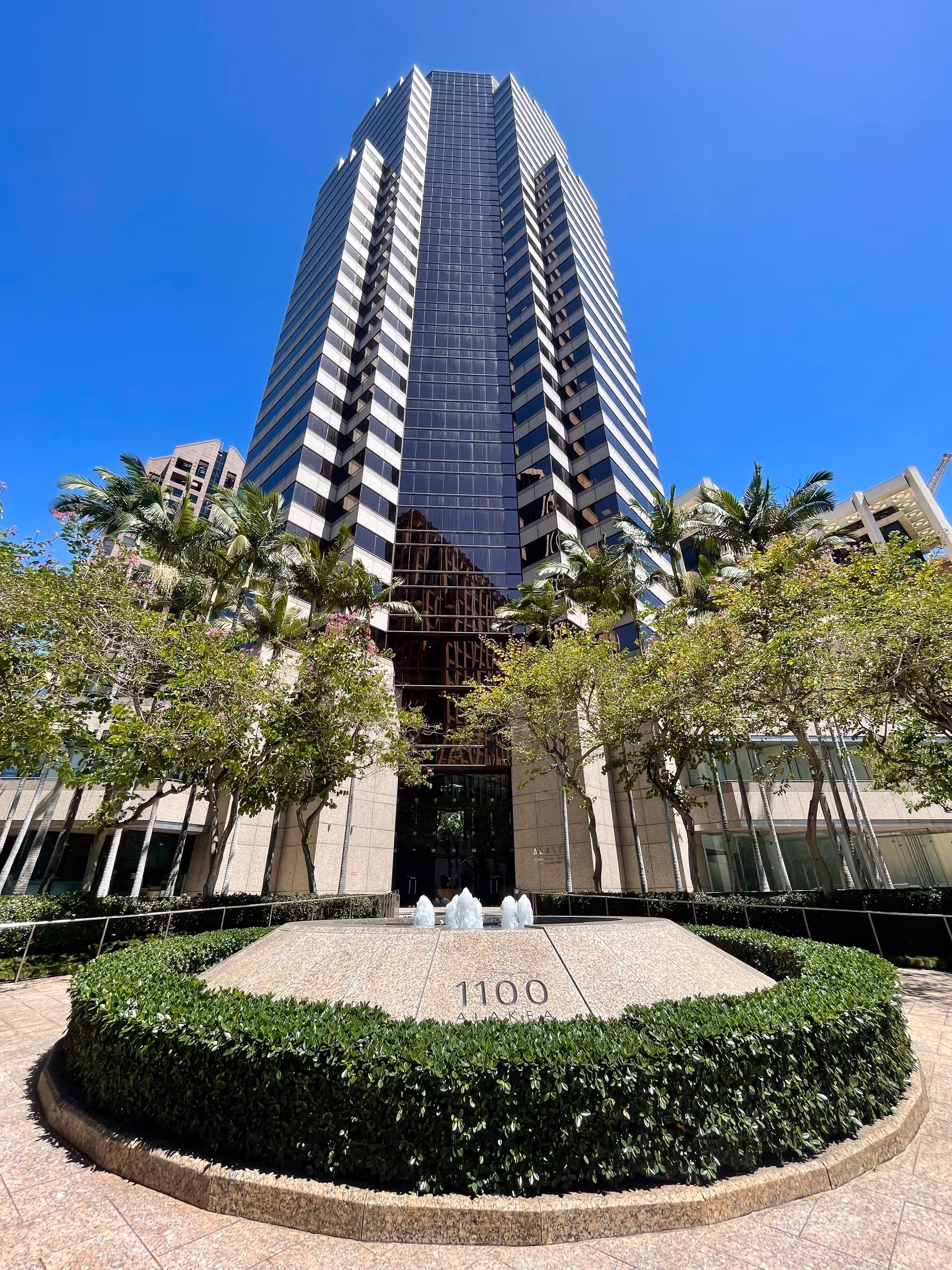 Tall glass skyscraper with a circular fountain, hedges, and a clear blue sky