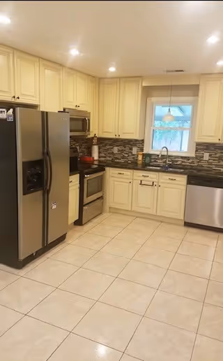 Bright kitchen with cream cabinets, stainless steel refrigerator, stove and dishwasher, tiled floor and a window over the sink.