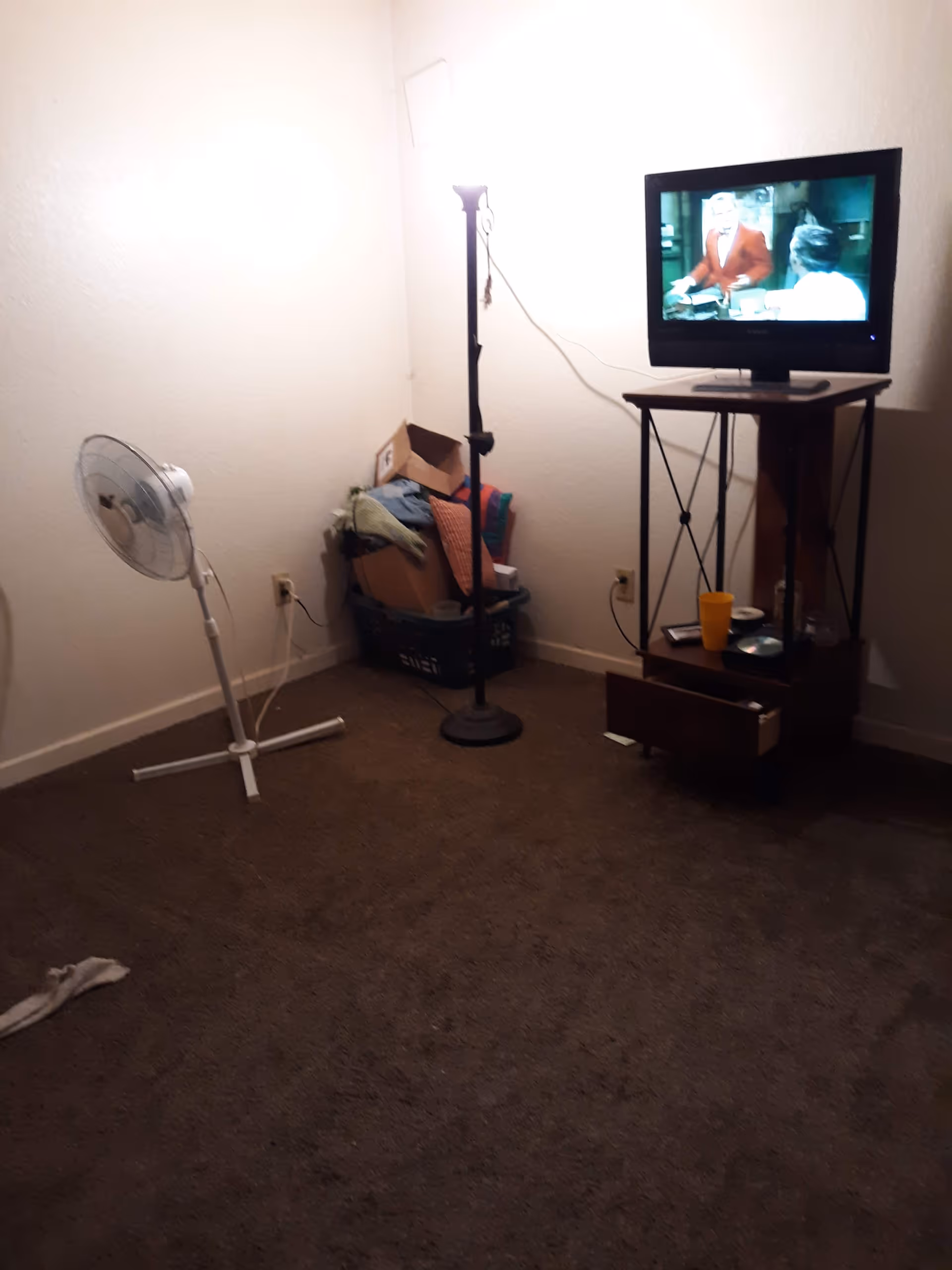 Small furnished room with TV on a stand, floor fan, and a cluttered laundry basket.