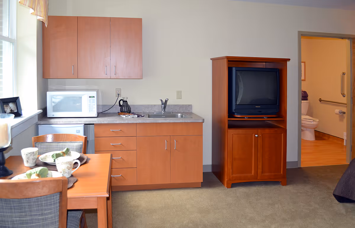 Interior view of a senior living facility room featuring a small kitchenette with wooden cabinets, a microwave, a sink, and a coffee pot. To the right, there is a wooden cabinet with an older model television. A dining table with two chairs is partially visible in the foreground, set with cups and plates. In the background, an open door reveals a bathroom with a toilet and grab bars.