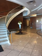 A modern lobby with a curved staircase, glass railing, and a vase on a pedestal.