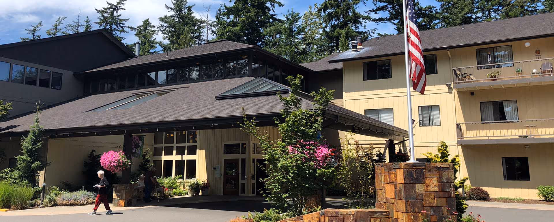 Exterior view of a multi-story senior living facility building with beige siding and a dark roof. There is a covered entrance with large windows, landscaping with bushes and flowers, an American flag on a flagpole, and two people walking near the entrance. Tall trees are visible in the background under a partly cloudy sky.