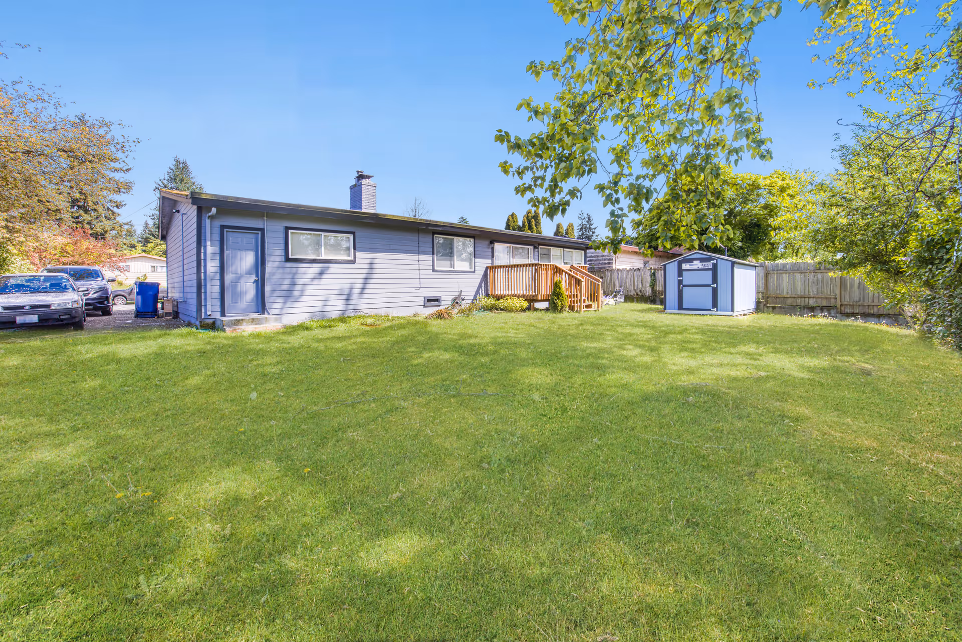 A single-story blue house with a wooden deck, shed, and green lawn.