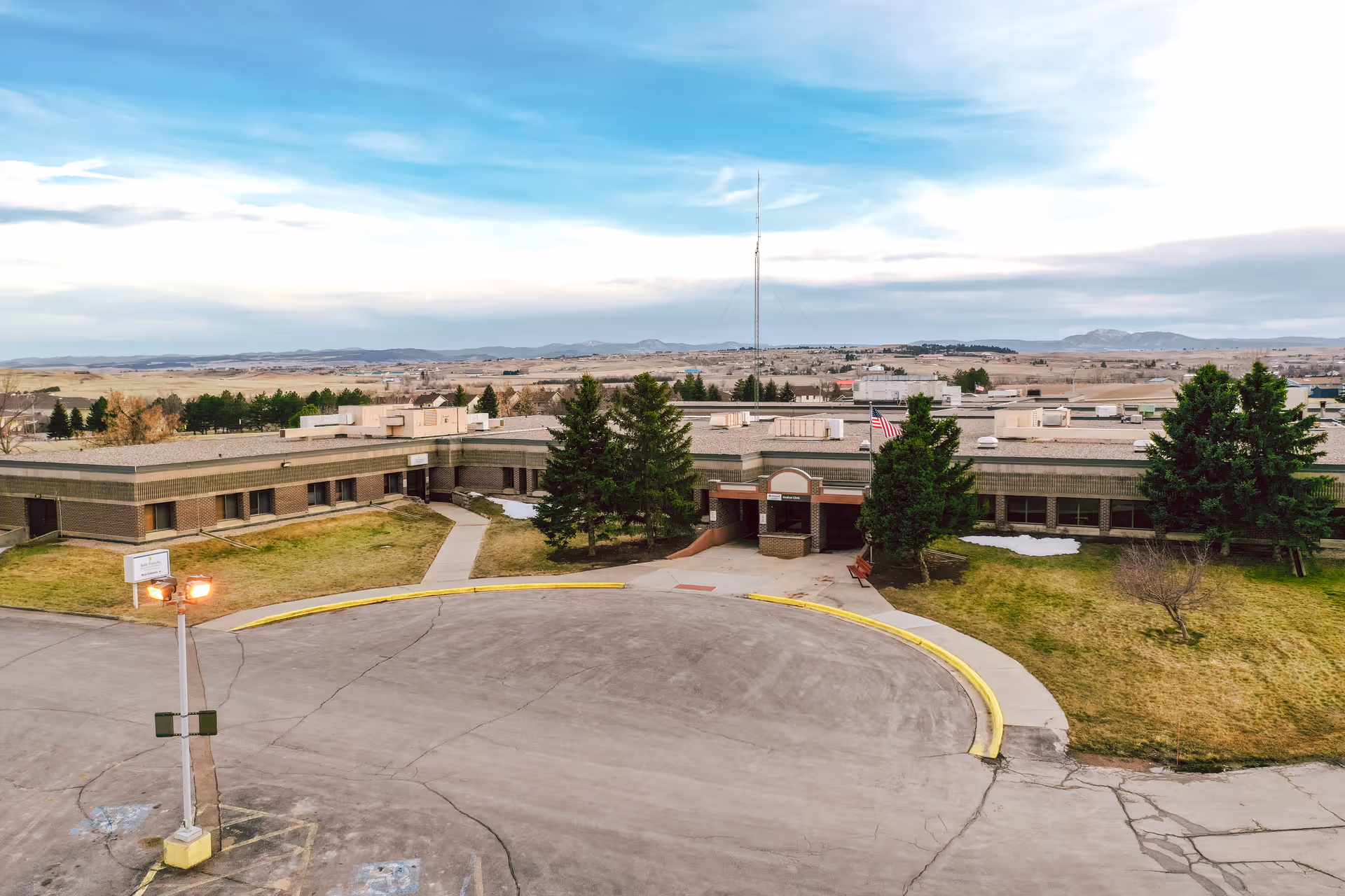 Aerial view of a single-story healthcare facility building with a circular driveway in front, surrounded by grass and trees, under a partly cloudy sky with distant hills in the background.