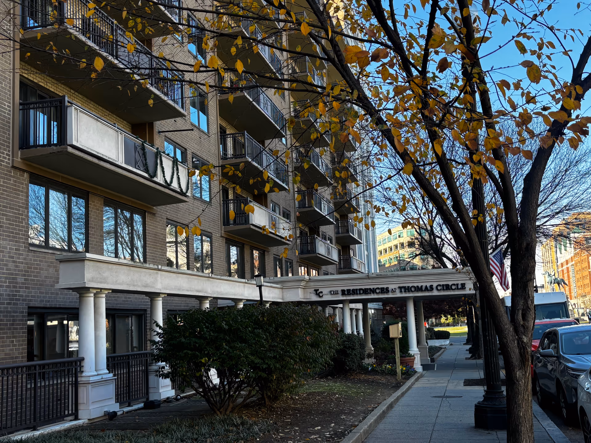 Multi-story brick residential building with balconies, a columned entrance canopy, trees with yellow autumn leaves, and cars parked along the sidewalk.