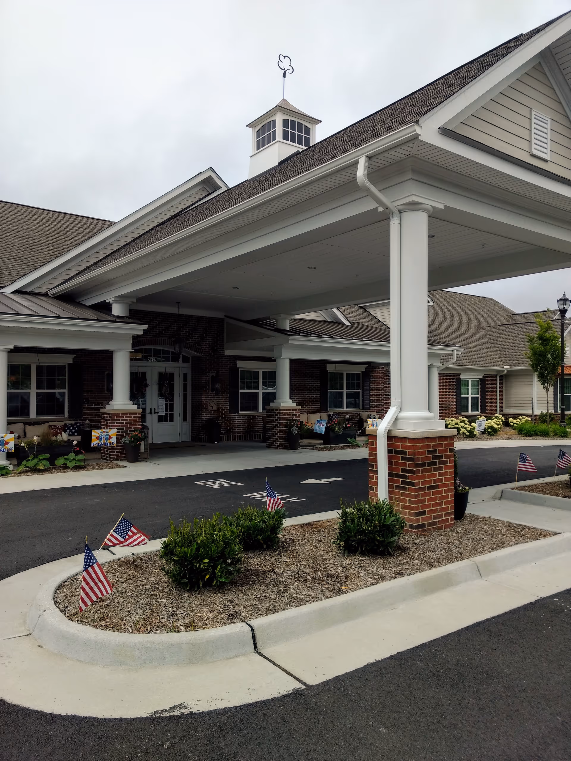 Covered entrance of a senior living facility with brick columns and flag-decorated landscaping.