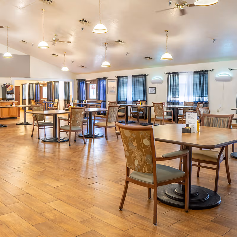 A spacious senior living dining area with multiple tables, chairs, pendant lights, and blue-curtain windows.