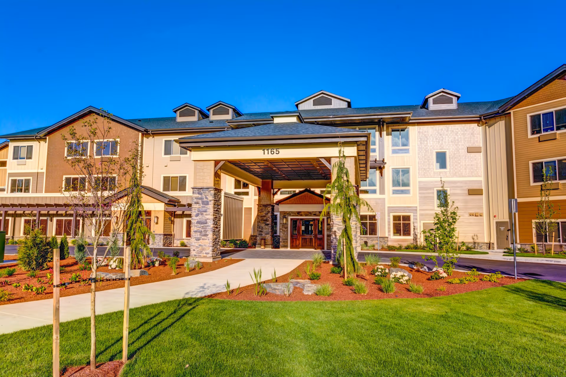 Front exterior view of a senior living facility named Village at Keizer Ridge with a covered entrance, stone pillars, well-maintained landscaping, and a clear blue sky.