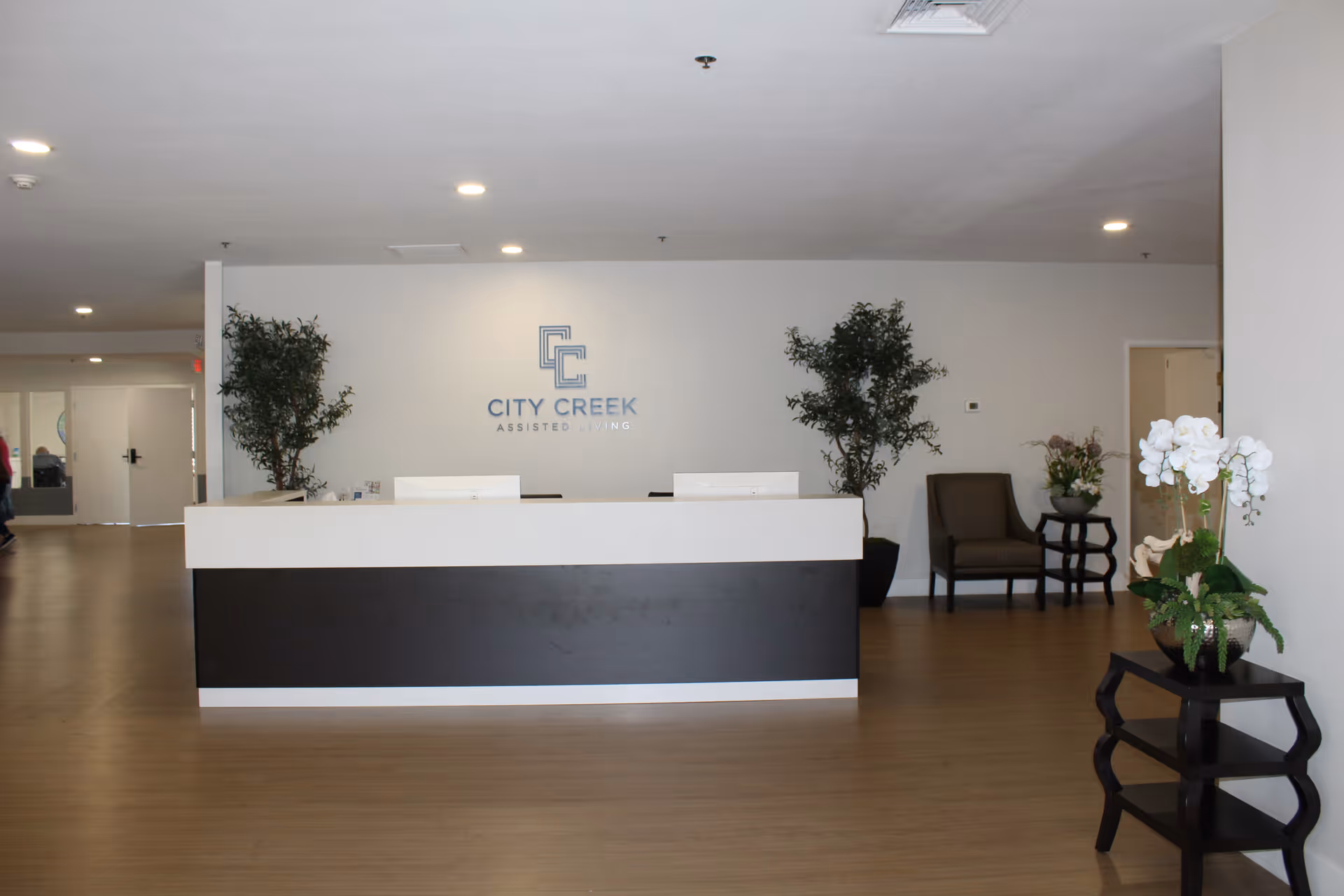 Reception area of City Creek Assisted Living facility with a modern front desk, two potted plants behind the desk, a chair and side table with flowers to the right, and a small table with white orchids in the foreground on the right side.