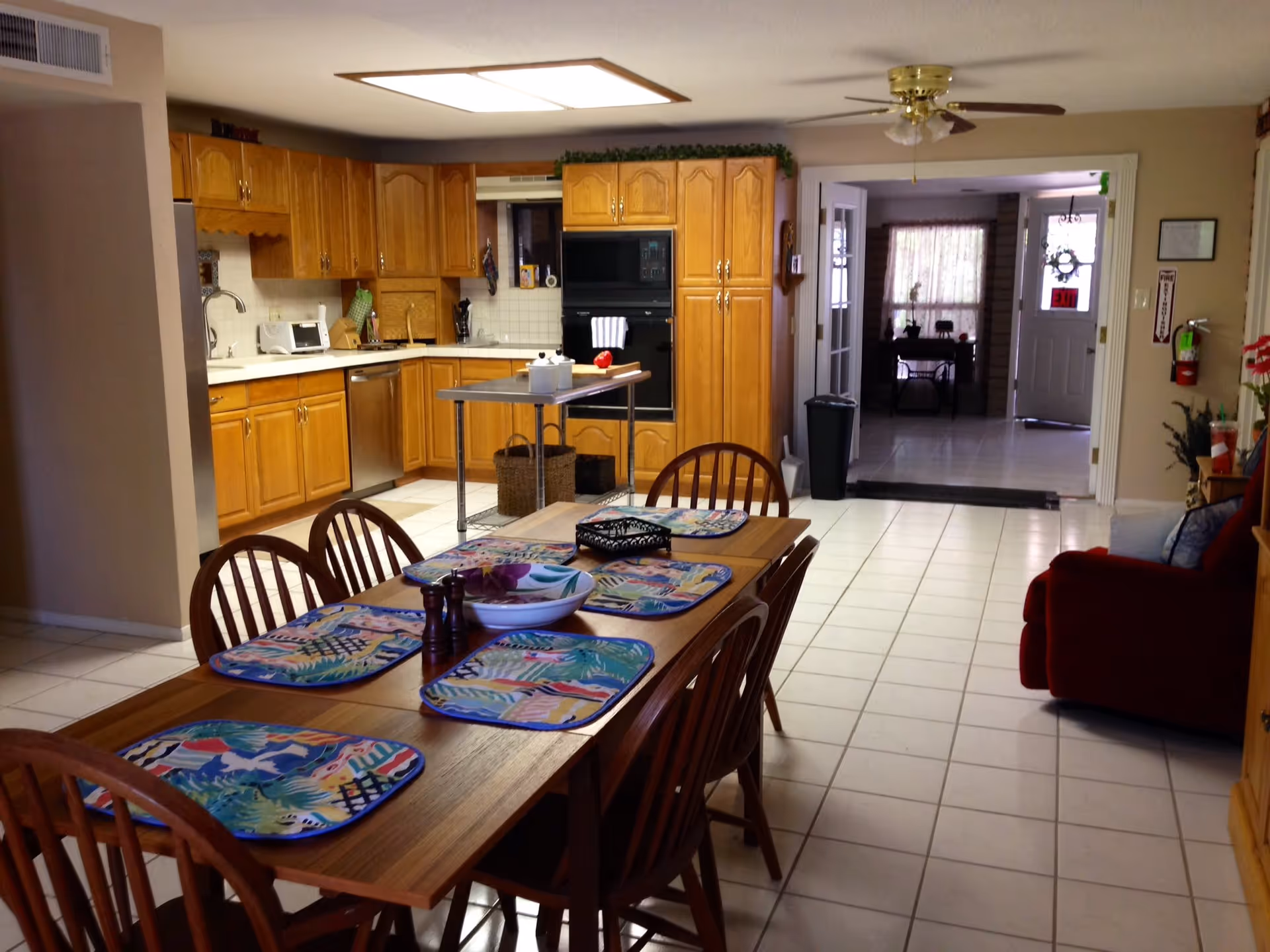 A spacious kitchen and dining area with wooden cabinets, a stainless steel dishwasher, and a wooden dining table set with six chairs and colorful placemats. The kitchen has white tiled floors and a ceiling fan, with an open doorway leading to another room with a door marked EXIT.