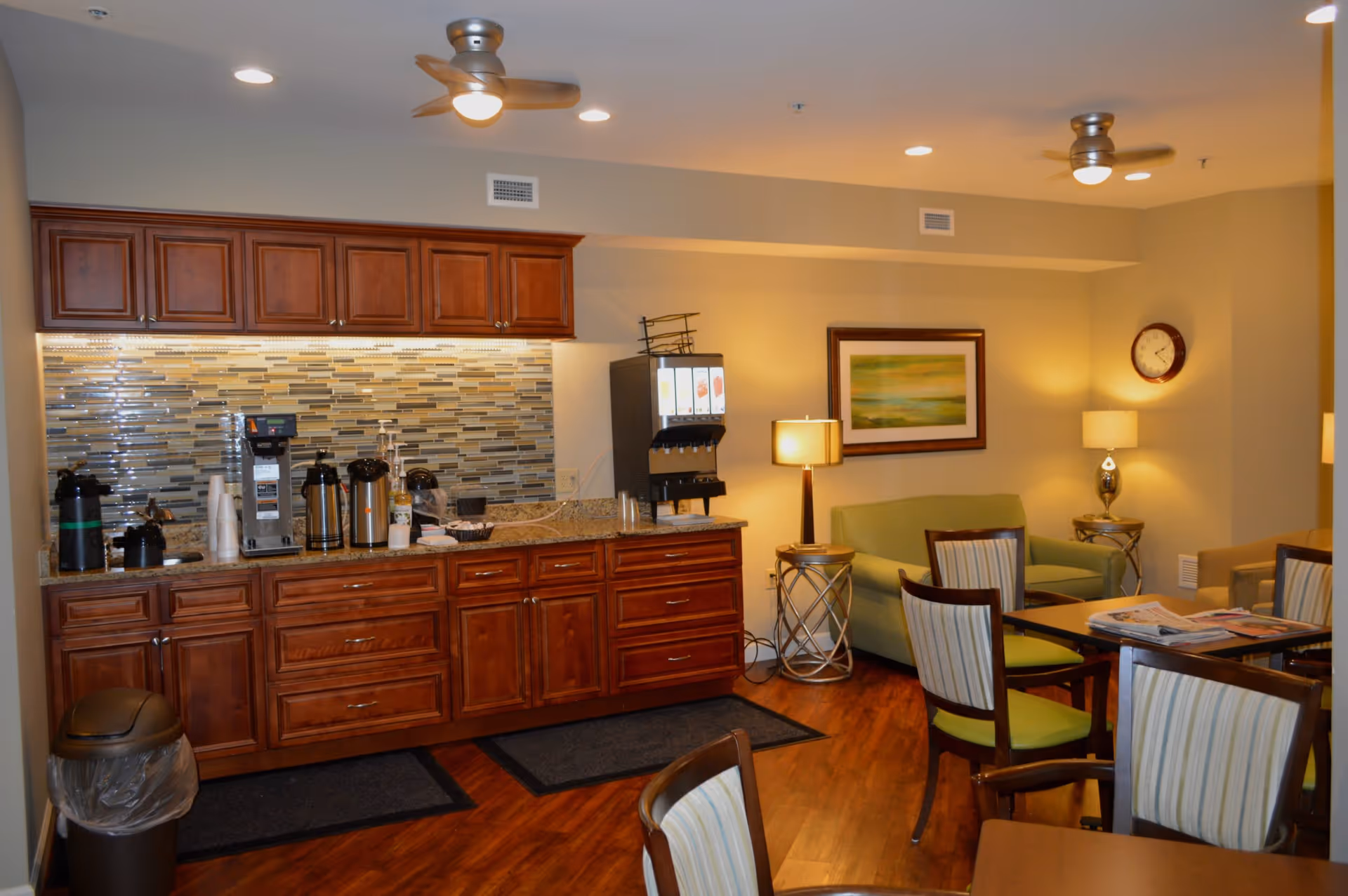 Interior view of a common area in a senior living facility featuring a coffee and beverage station with wooden cabinets and a tiled backsplash. The room includes seating with striped cushioned chairs, a green sofa, side tables with lamps, a wall clock, and framed artwork. The floor is wooden, and ceiling fans with lights are visible.